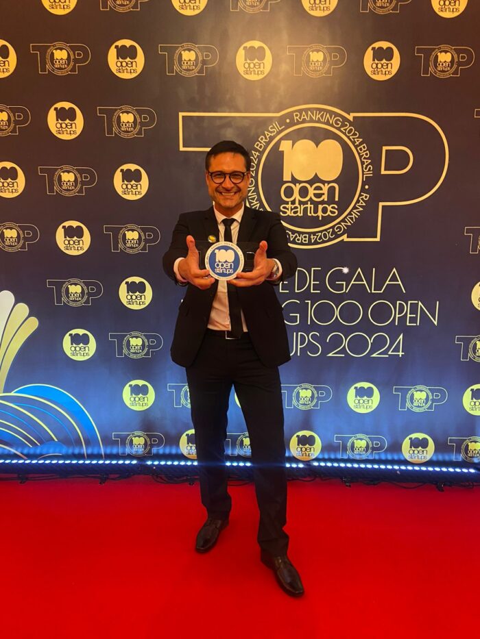 Homem sorridente, usando terno e gravata, segura um troféu com o logotipo '100 Open Startups' em destaque. Ele está em um tapete vermelho, posicionado em frente a um banner que exibe várias vezes o logotipo '100 Open Startups' e a frase 'Ranking 2024 Brasil.'